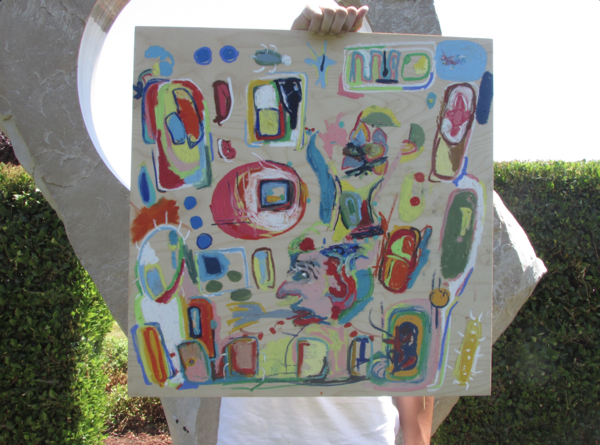 A person holding up a large painting outdoors: bright red, yellow, blue and green shapes with a face in profile, on a pale wooden panel.